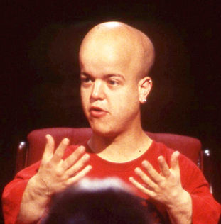 Tom Shakespeare wearing a red shirt gesturing with both hands as if talking to someone beyond the camera.