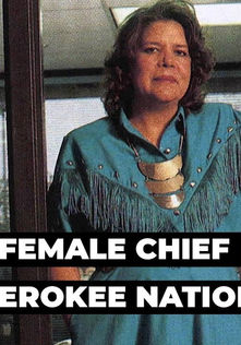 Chief Wilma Pearl Mankiller with shoulder-length wavy brown hair. She wears a teal fringe dress with brass studs and a necklace of three metallic half circles.