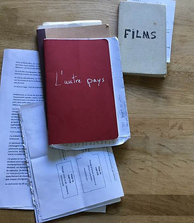 plusieurs carnets et des feuilles volantes posées sur une table. Deux carnets sont titrés, l'un "L'autre pays", le second "FILMS"