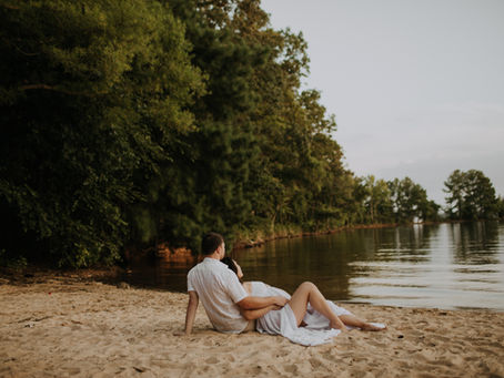 A husband and wife sitting on the beach looking at the water for their Lake Norman maternity session