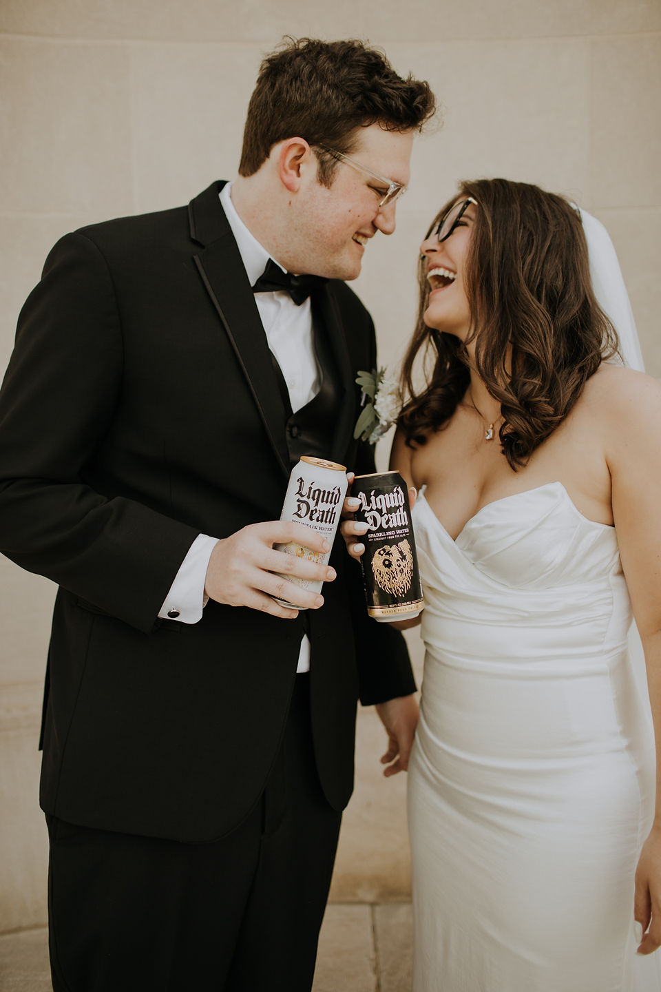 A bride and groom both holding liquid death water laughing at each other