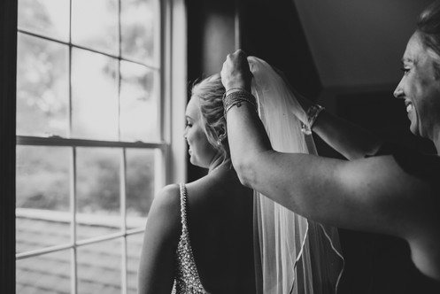 a woman putting a veil in a bride's hair while she looks out a window for her charlotte micro wedding photographer