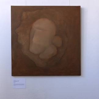 Na ścianie wiszą trzy obrazy Krystiana Lichonia; w centrum „Nie jesteś sam” (80 × 80 cm) – ciepłobrązowe pole, w którym miękko modelowana, zaokrąglona bryła z kilku przylegających do siebie owalnych segmentów wyłania się z ciemniejszego „zagłębienia”. Po lewej płótno w jaśniejszych beżach z mgiełkową, owalną plamą; po prawej ciemniejsze, oliwkowo‑brązowe tło z różowo‑łososiową, segmentową formą i wąskim, poziomym pasem zieleni u dołu.