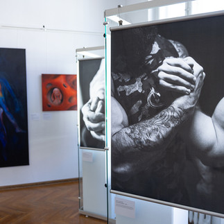 Na pierwszym planie widoczna jest fotografia Lidii Misiuny: czarno‑białe ujęcie przedstawiające skrzyżowane, zaciśnięte przedramiona i dłonie, z detalem tatuażu na skórze; w tle znajdują się obrazy Pauliny Wojewodzic — po lewej pionowe płótno o ciemnej tonacji z abstrakcyjnie ujętą sylwetą rozświetloną barwnymi wyłomami, po prawej kwadratowa kompozycja w odcieniach czerwieni i pomarańczu, przypominająca perforowaną, miękką materię z osadzoną w jednym z otworów owalną, organiczną formą przypominającą zrolowane ciało.