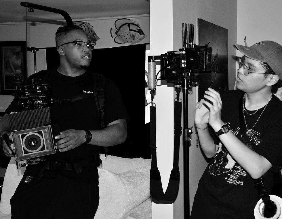 Two people in a room handle camera equipment. One holds a large camera; the other adjusts a rig. Wall decor includes fish figures. B&W image.
