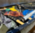 Fly-fishing gear laid out in a canoe on calm water, including a rod, net, leather case, and an Orvis tube in bright sun