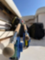 Fishing rods with blue reels and cork handles on a boat deck with bags. Bright sky overhead, suggesting a calm, sunny day.