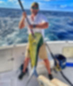 Man proudly holds large mahi-mahi on a boat, ocean behind him. He's wearing a cap and sunglasses, smiling. Sky is clear and blue.