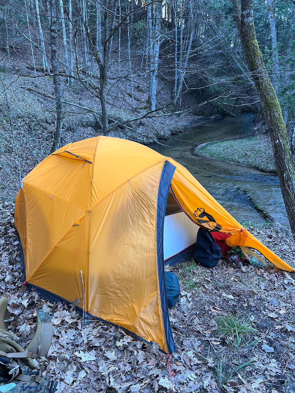 Orange/yellow tent pitched by a river in the woods.