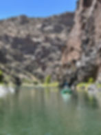 Two people raft a green inflatable boat on a calm river between steep rocky canyon walls under a clear blue sky