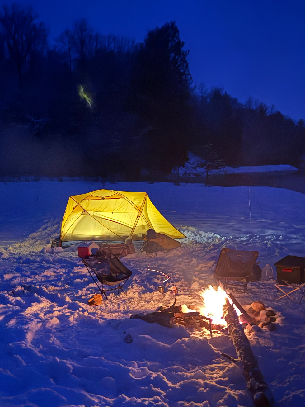 Yellow tent glowing in snowy campsite at night. Chairs and campfire nearby. Trees silhouette the background. Peaceful, cozy mood.