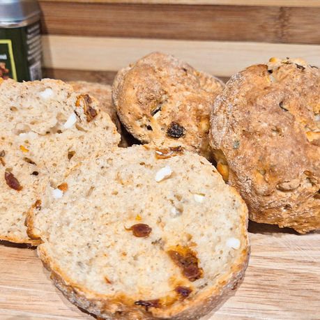 A freshly cooked batch of bagels with sun dried tomatoes and halloumi on a wooden board beside a green tin labeled "OliveOlive Greek Seasoning Blend" for Souvlaki. Rustic and appetizing.