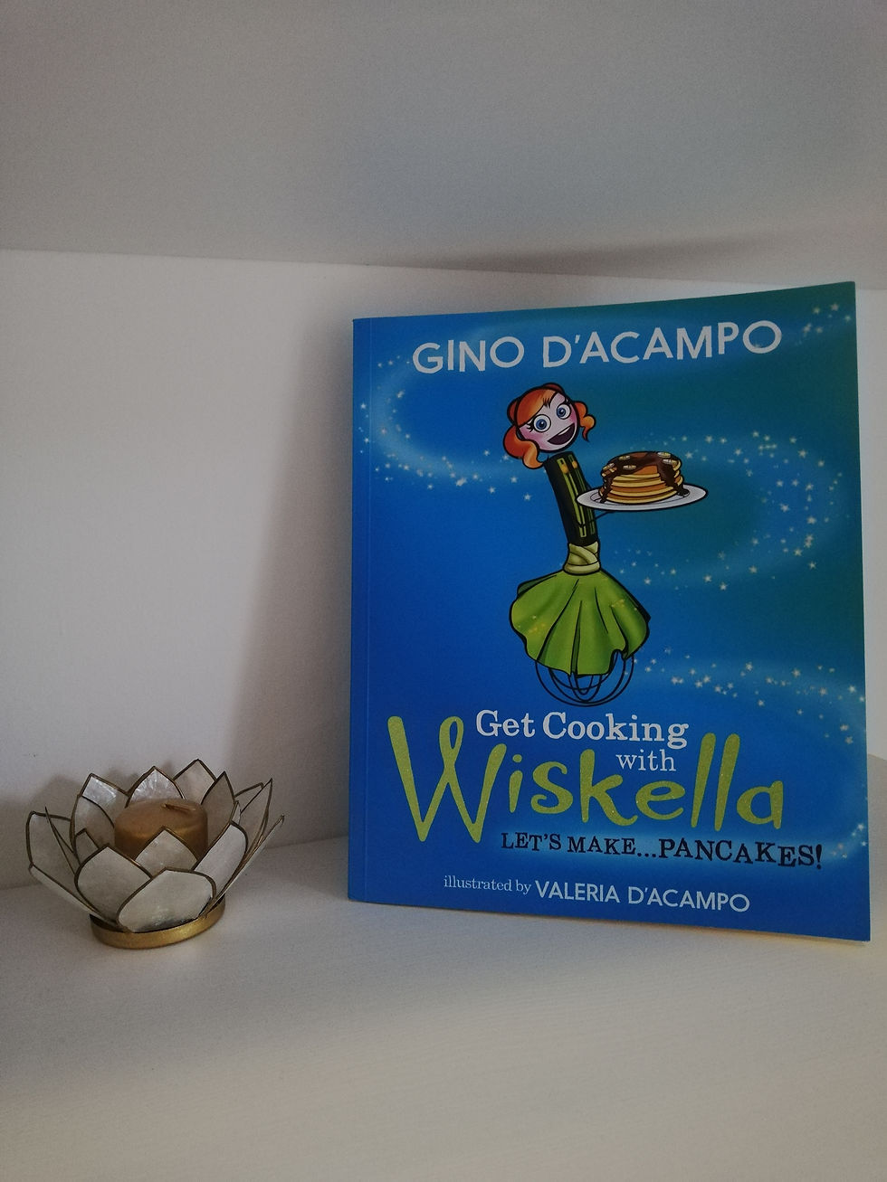 la copertina è blu, si vede il nome dell autore Gino D'acampo e l'immagine di un frustino da cucina umanizzato, vestito da donna, con capelli rossi, che porta in mano un piatto di pancake