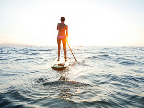 Person paddleboarding alone on calm ocean water at sunset, moving toward the horizon with warm sunlight reflecting across the waves.