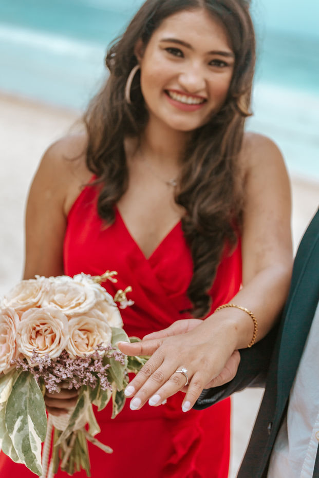 A beaming woman holds up her left hand, showing off her engagement ring to the camera with pure joy and excitement. The ring sparkles in the light, with a stunning diamond nestled on a delicate gold band. Her smile is infectious and her happiness radiates through the photo. The background is blurred, allowing all attention to focus on the ring and the woman's expression. It's a moment of pure happiness, captured in time forever.