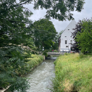 Ein kleiner Bach fließt durch grüne Wiesen und im Hintergrund eine kleine Brücke mit weißem Haus.