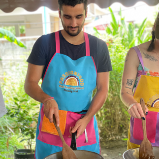 Ein junger Mann mit einer knallbunten Schürze steht an einem Wok und rührt mit einem Holzrührer das angebratene Essen.