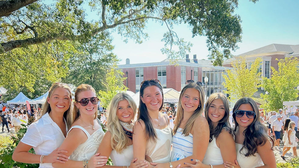 Seven smiling women in summer dresses pose outdoors under trees. Background shows a crowd with tents and a brick building. Sunny day.
