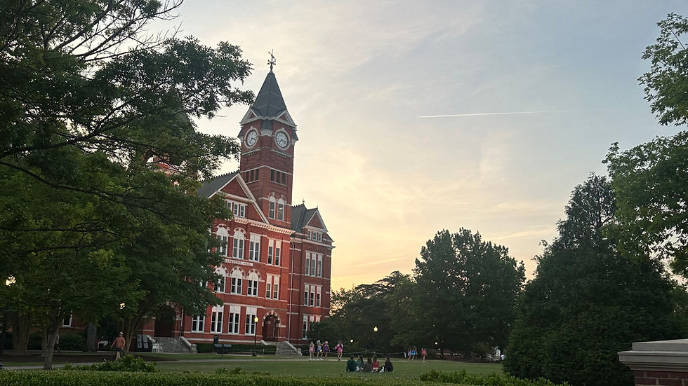 Red brick clock tower with "Auburn" sign, people walking and sitting on the lawn. Evening sky with trees framing the scene. Tranquil mood.
