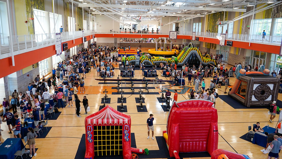 Indoor event with people enjoying inflatables in a gym. Red and yellow slides, booths, and exercise bikes. Bright, lively atmosphere.