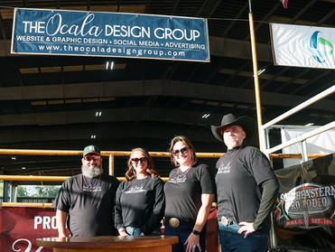Four people in black shirts stand under a blue "The Ocala Design Group" banner in an indoor rodeo setting, smiling confidently.