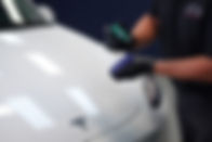 A close-up of Bill preparing to apply a ceramic coating, carefully pouring Gyeon Base Type 2 professional coating onto a microfiber applicator block with a white Tesla in the background.