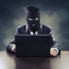 Man in a suit wearing a black ski mask sits at a desk using a laptop, symbolizing online scams and cybercrime.