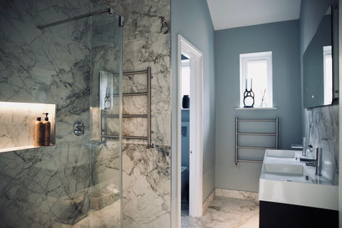 A dark marbled walk in shower stands to the left of this modern bathroom loft conversion while to the right stand white rectangular his and her sinks. Behind is a doorway and window leading to other parts of the loft conversion.
