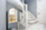 An elegant traditional staircase installed in a loft conversion used as the main image for The Loft Build Company with grey walls, beige carpet, white wood and a arched frosted window behind.