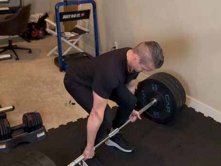 A person lifts a heavy barbell in a home gym. Background includes a "Just Do It" sign, a punching bag, and exercise equipment. Mood is focused.