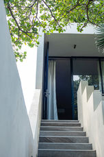 A straight-on view of an outdoor concrete staircase leading up to an open dark doorway framed by clean white stucco walls, white curtains, and overhanging green tree leaves.