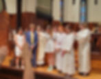 People stand together at St. Mark's Episcopal Church during a blessing ceremony. Some wear robes, others formal attire. Stained glass windows in background.