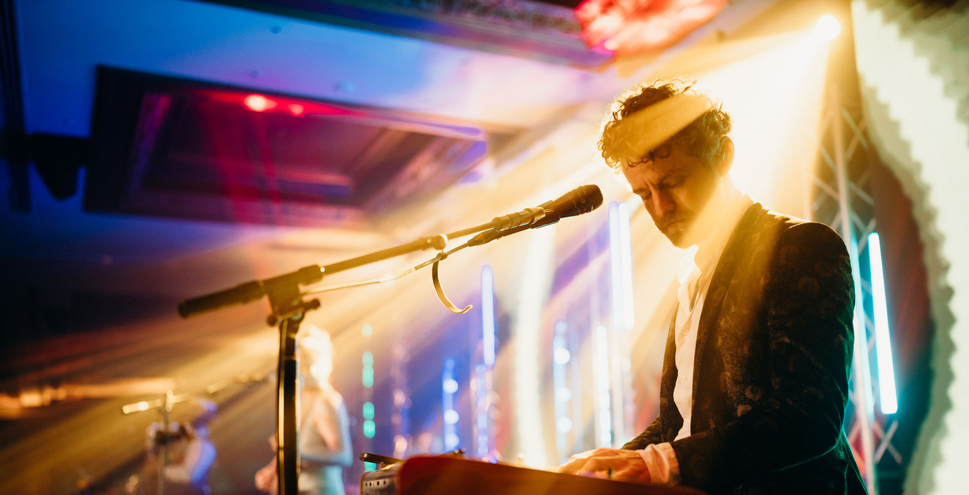 A man playing the keyboard, with concert lighting shining behind him and an LED screen in the background.