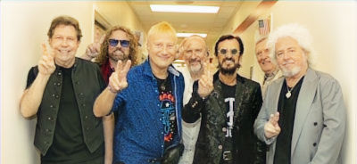 Six band members of Ringo Starr's All-Starr Band in a hallway, smiling and flashing peace signs; black-and-white group portrait with a relaxed, cheerful mood.