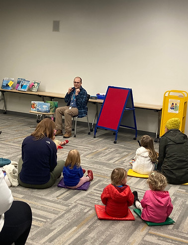 Man reading to children. Red easel and books on a table. Canby Friends Of The.