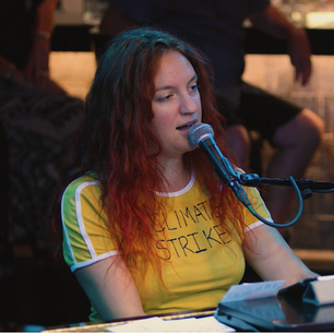 [image description: Elizabeth sits at the piano, singing into a microphone, at a crowded bar. Her hair is red, and she wears a t-shirt that says #climatestrike.]