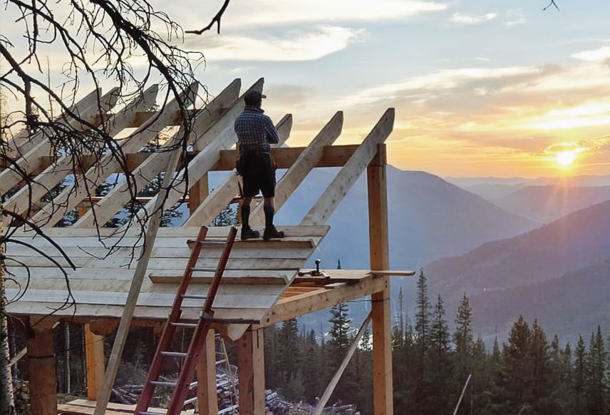 A craftsman stand on top of a timberframe building looking out at the sunset.
