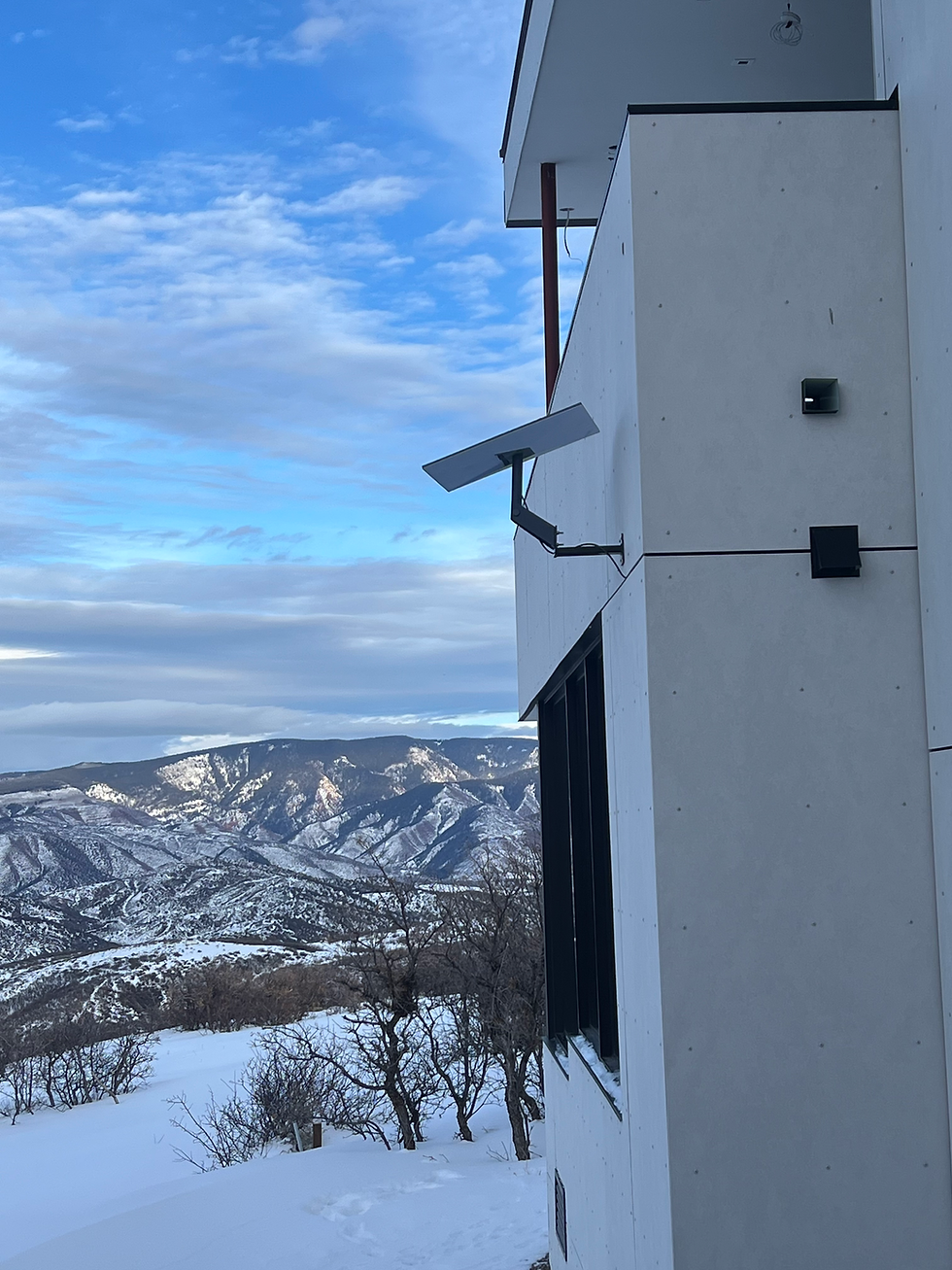 A Starlink dish mounted on a custom-fabricated bracket extending from the exterior wall of a modern home, ensuring proper clearance. Snow-covered mountains and a blue sky with scattered clouds create a scenic backdrop.