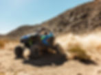A blue off-road vehicle with "RZR" text speeds through a rocky desert, kicking up dust. Clear blue sky and rugged terrain in the background.