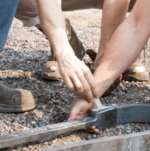 Two hands reach down to hold a forged piece of metal. One hand holds chalk to mark the metal