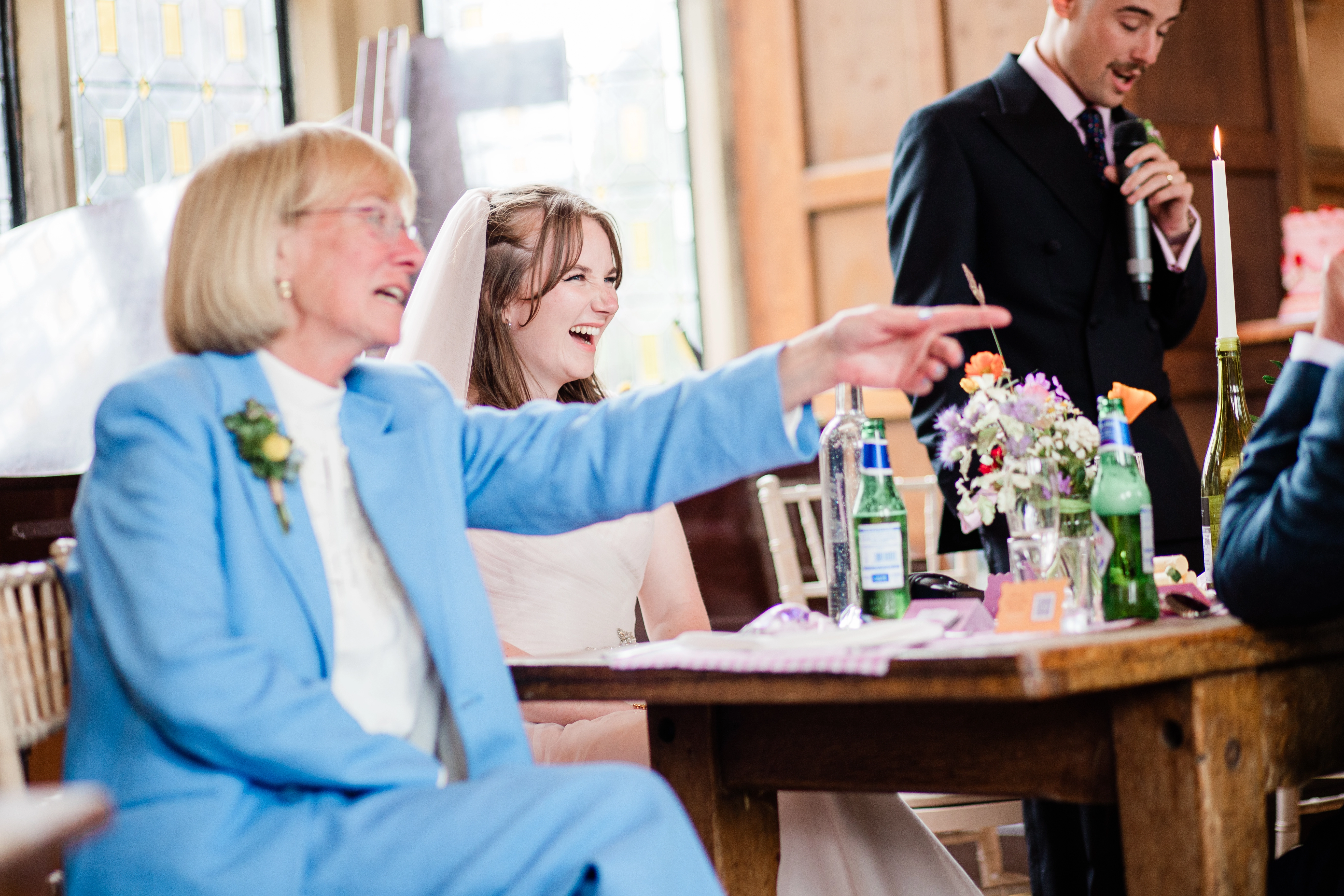 The bride laughing at the grooms speech while her mum points at someone off camera