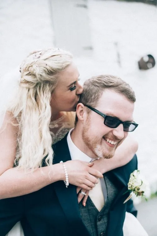 A bride on the back of a groom giving him a kiss on the back of the head