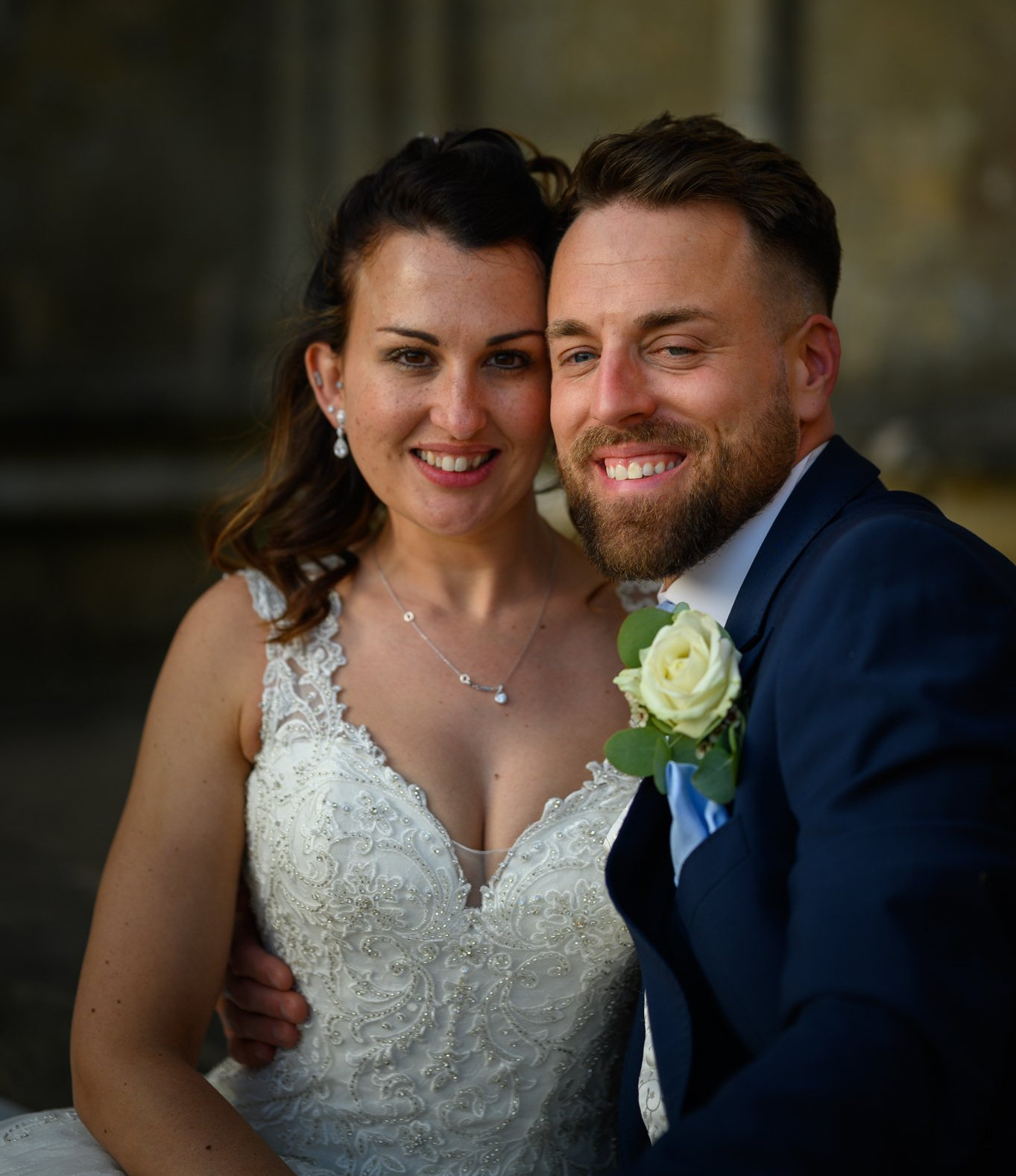 Smiling bride in a beaded white gown and groom in a navy suit with a white rose pose close together outside at Newstead abbey Wedding Venue.