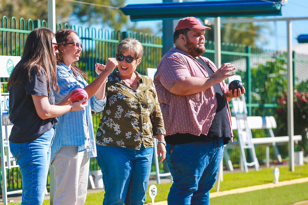 Four people joyfully playing lawn bowls outside; two hold bowls, one cheers. Sunny day with green fencing and blue skies.