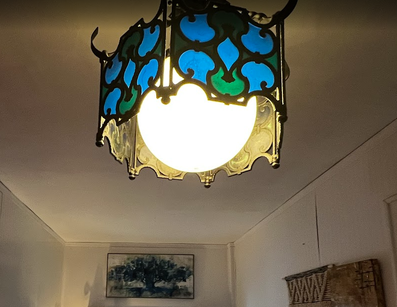 Ornate ceiling lamp with blue and green stained glass in a room with white walls. A framed picture and patterned wall decor are visible.