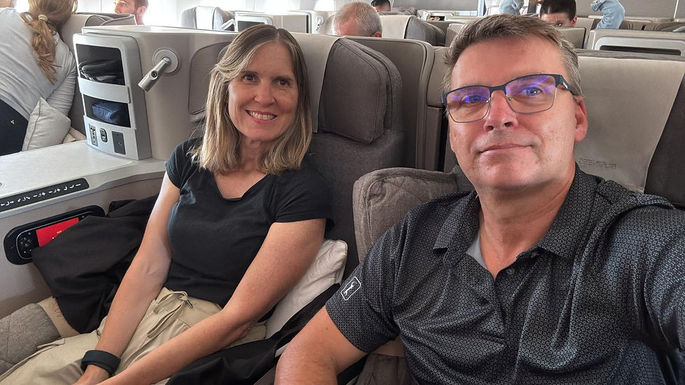Smiling couple taking a selfie in an airplane business-class cabin with beige seats and other passengers behind them.