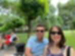 Smiling couple takes a selfie on a park bench, with leafy trees, grass, and other people sitting nearby in the background.