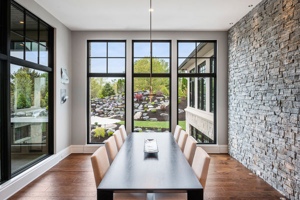 A modern dining room with a large black table and beige chairs, framed by floor-to-ceiling windows overlooking a landscaped garden with stone features and greenery, complemented by a stone accent wall.