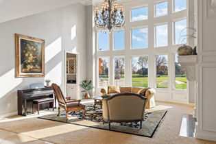 A bright and elegant living room with a large chandelier, grand windows overlooking a golf course, classic furniture, a black piano, and ornate architectural details.