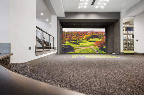 A luxury indoor golf simulator room with a high-definition screen showing a golf course, modern staircase railings, recessed lighting, and carpeted flooring.